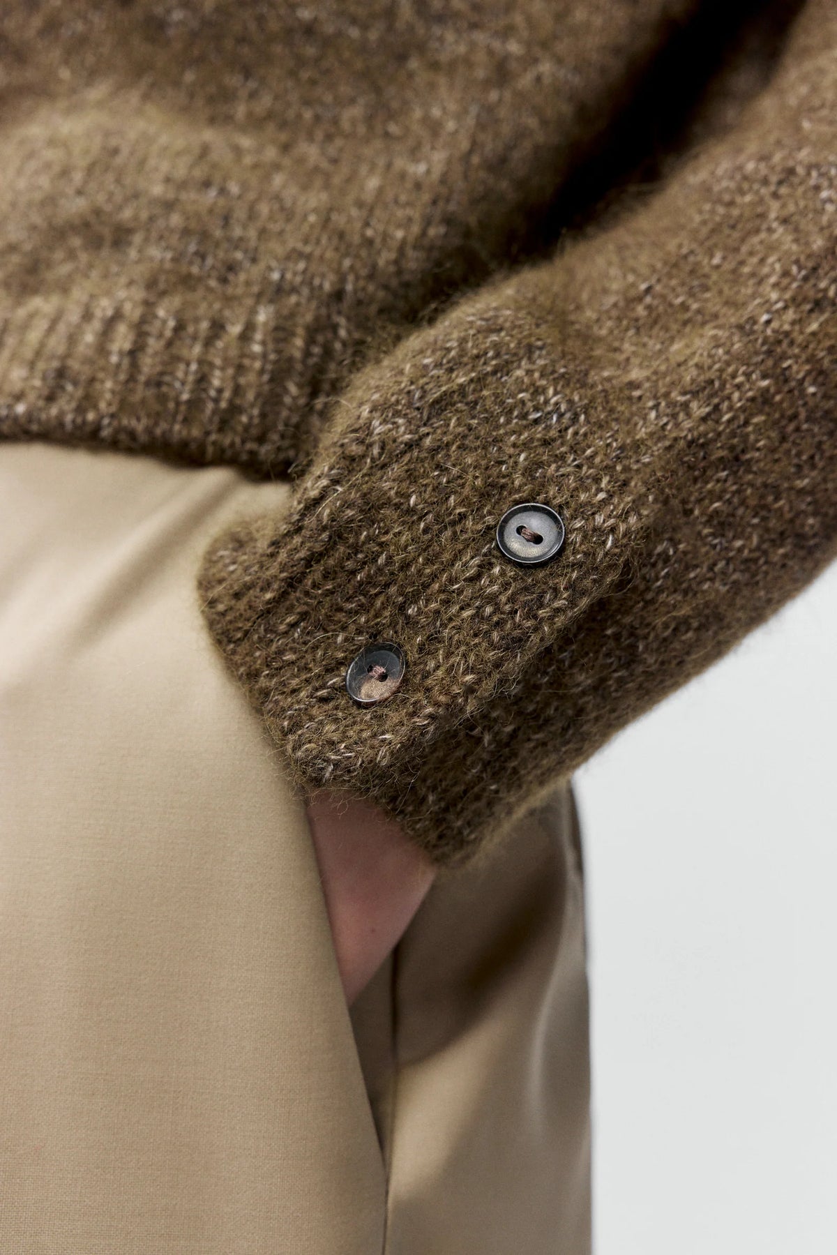 Close-up of a brown knitted sweater cuff with a button, worn over a beige garment.