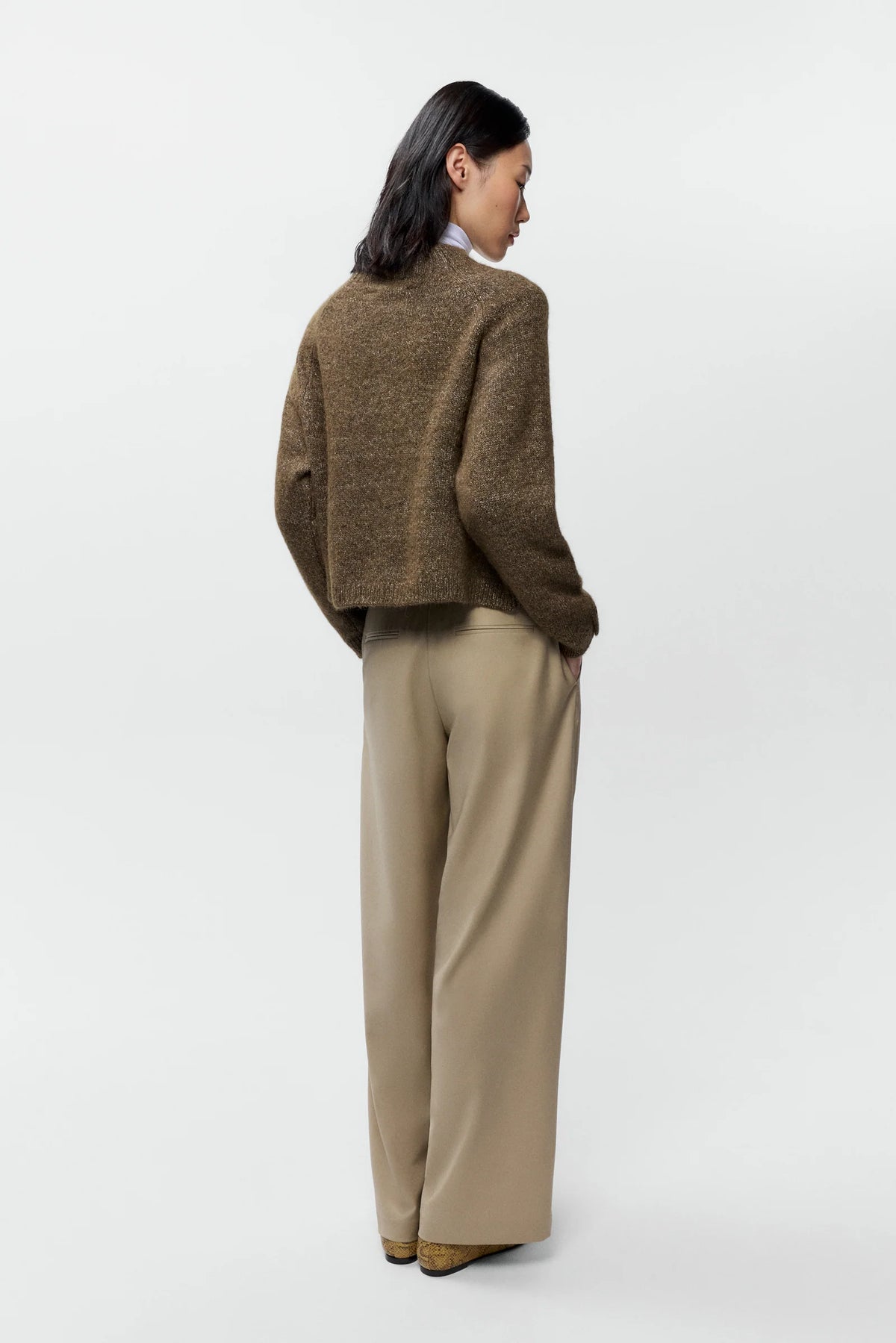 A model is shown from the back wearing a cropped, textured brown knit sweater and wide-leg beige trousers against a clean, light gray studio background.