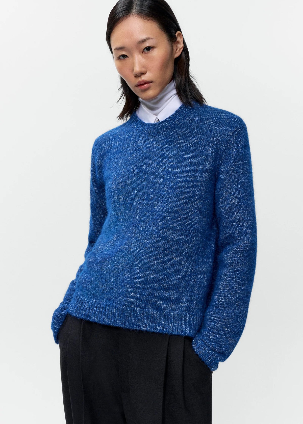 A model wearing a blue heathered knit crewneck sweater over a white collared shirt and black trousers, photographed against a clean white studio background.