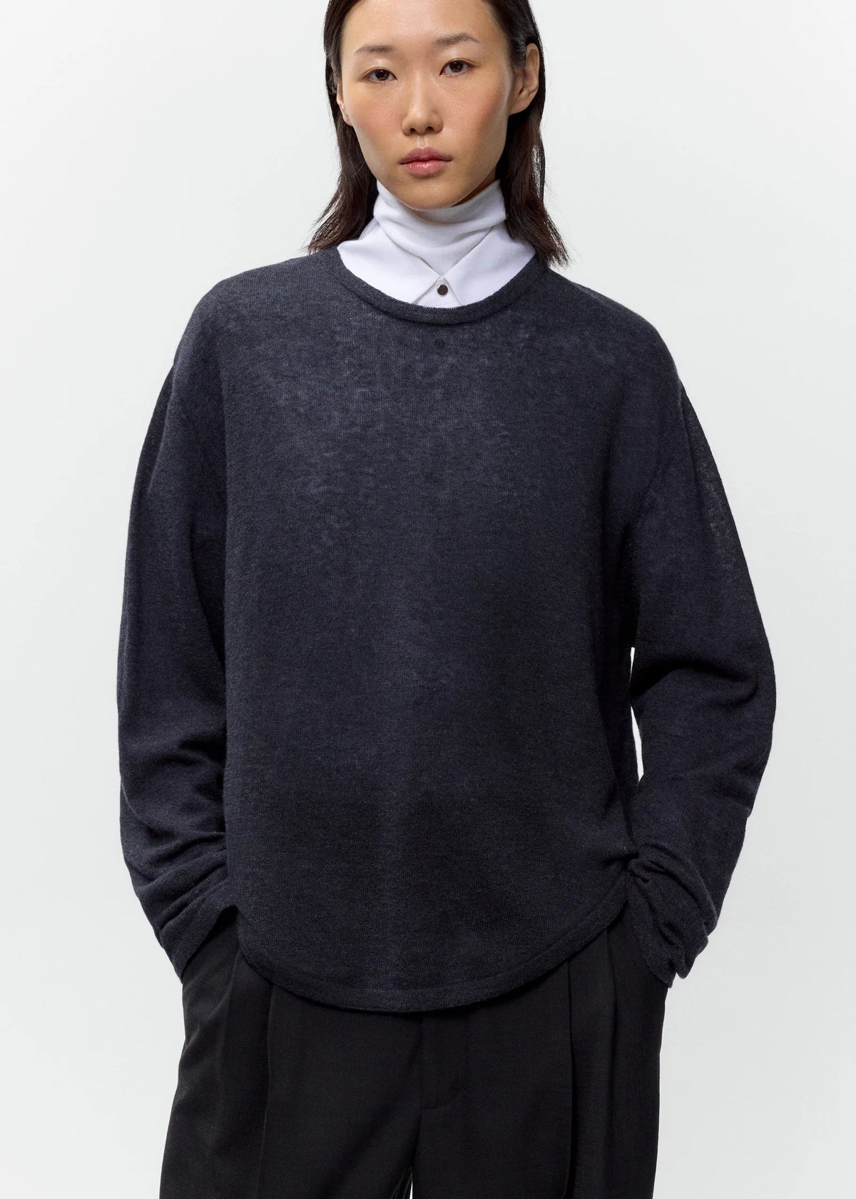 A model wearing a dark charcoal gray knit sweater layered over a white turtleneck and black trousers, photographed against a clean white studio background.