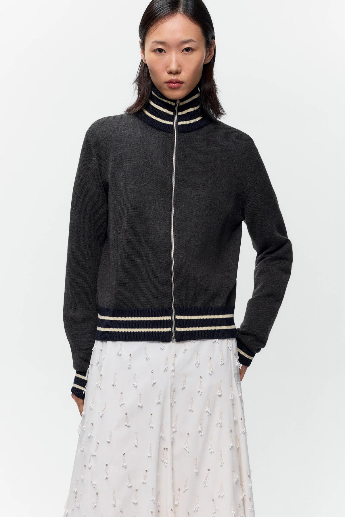 A model wearing a dark gray zip-up knit sweater with navy and beige striped ribbed collar, cuffs, and hem, paired with a white patterned skirt, against a plain light gray background.