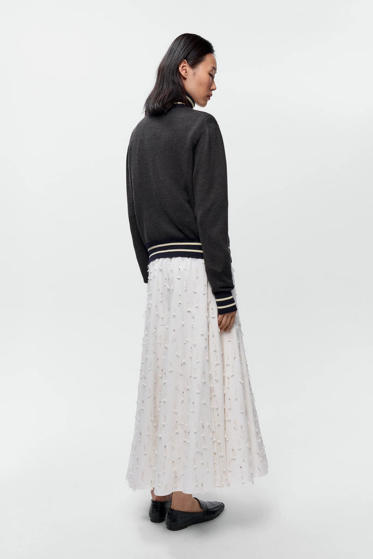 A woman is shown from the back wearing a dark charcoal gray knit cardigan with striped ribbed cuffs and hem, paired with a long white eyelet skirt and black flats, against a plain light gray studio background.