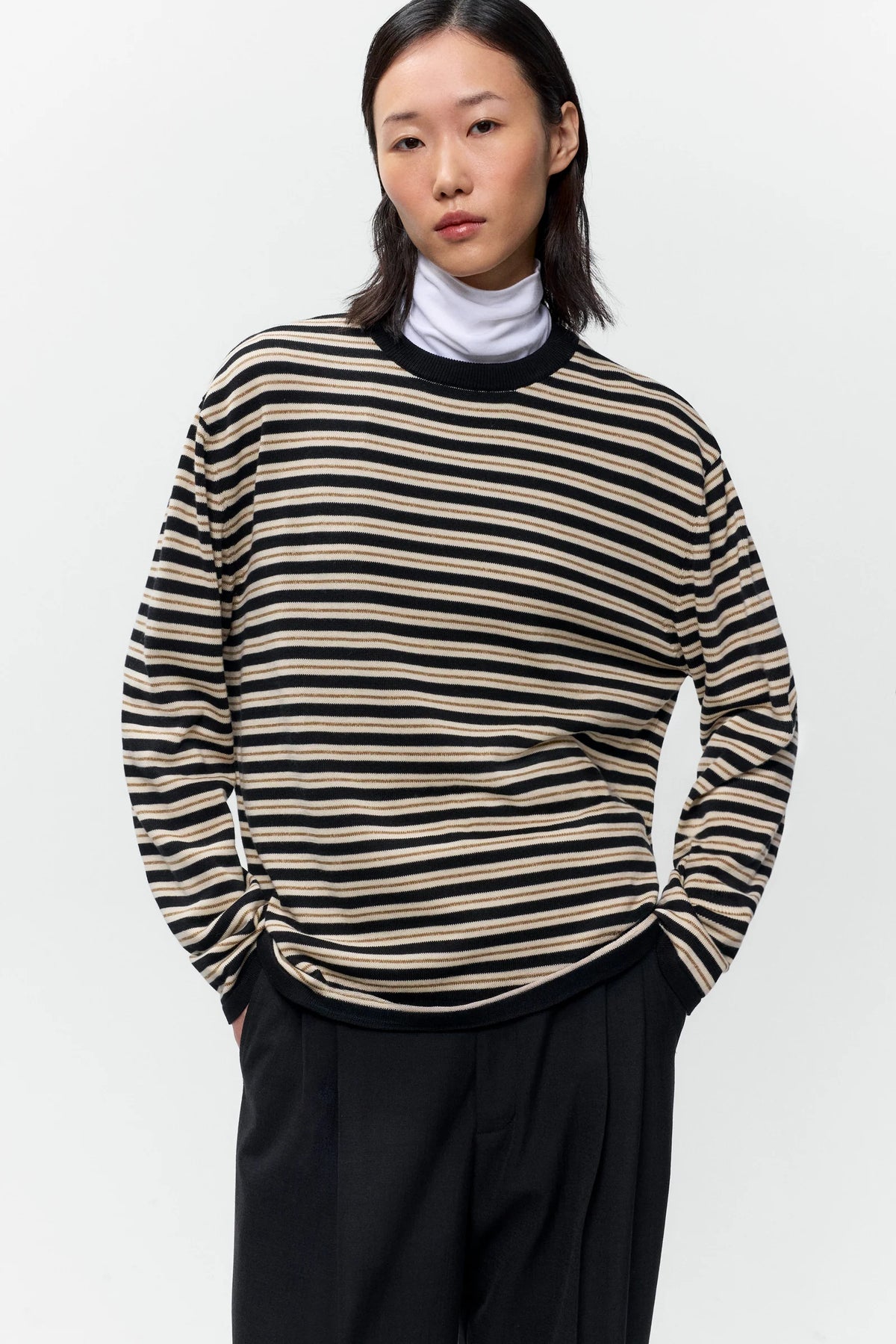 A model wearing a beige and black horizontally striped knit sweater over a white turtleneck, paired with black trousers, against a plain light gray background.