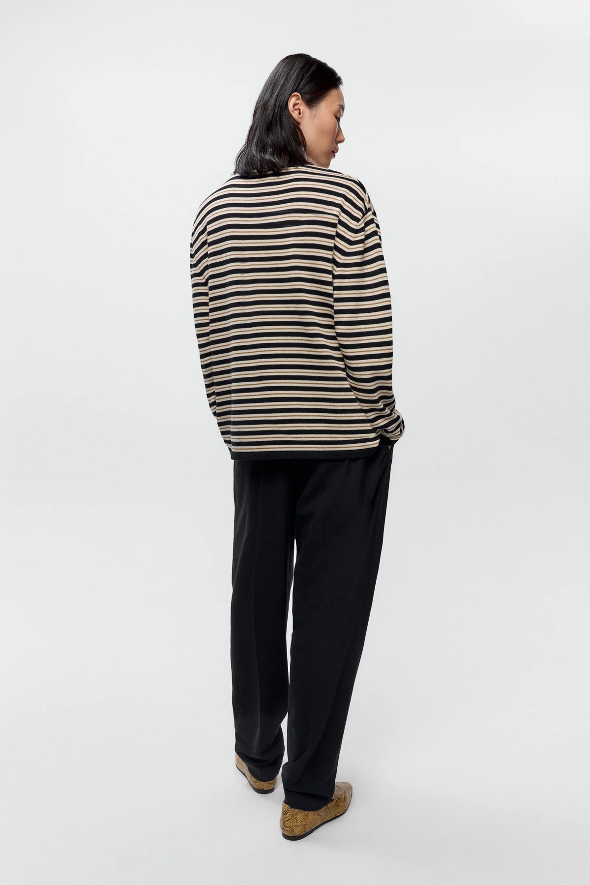 A model shown from the back wearing a beige and black horizontally striped long-sleeve knit sweater, black trousers, and tan loafers, set against a plain light gray studio background.