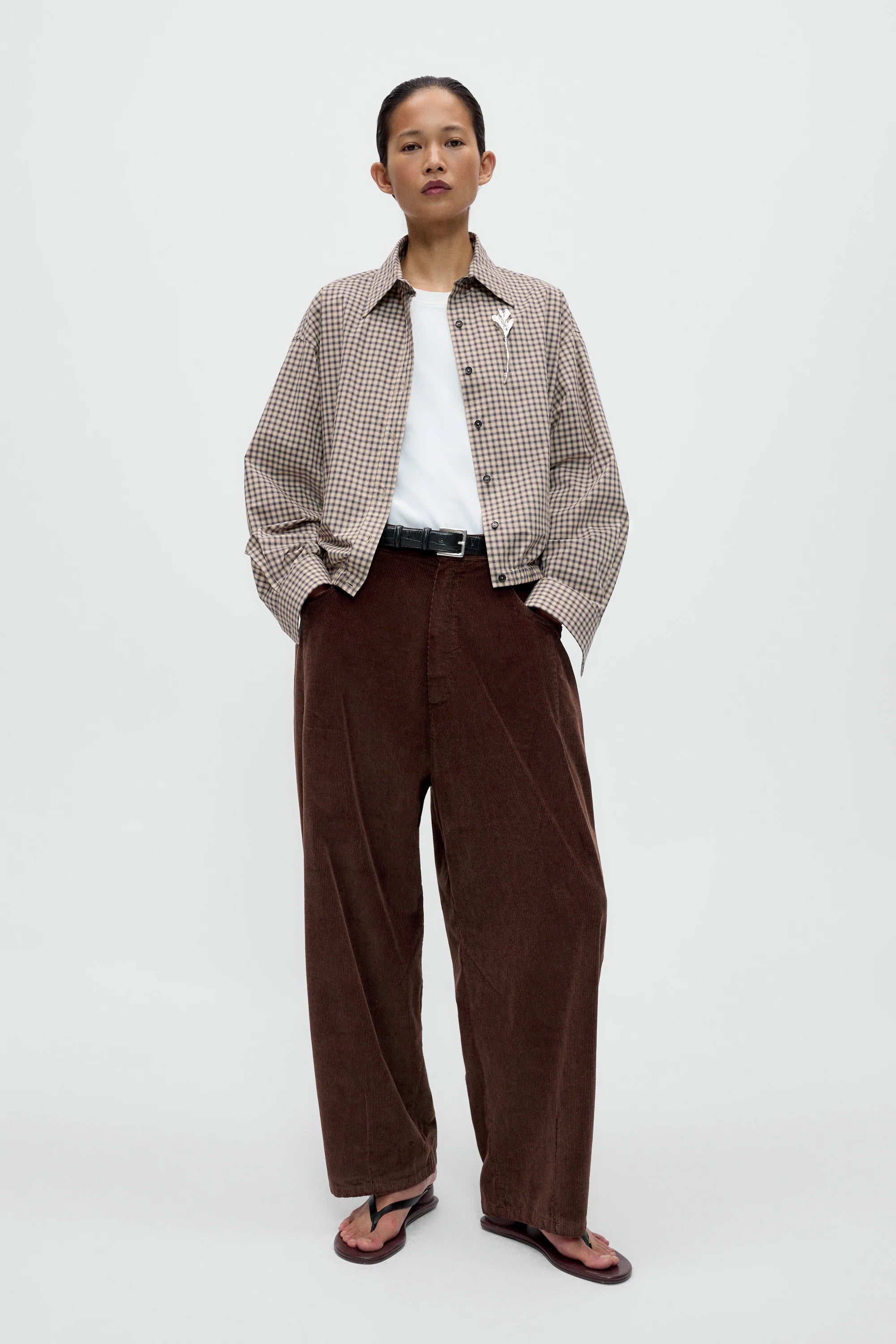 A model stands against a light gray studio background wearing a beige and brown checkered button-up shirt over a white t-shirt, paired with wide-leg dark brown corduroy trousers and dark sandals.