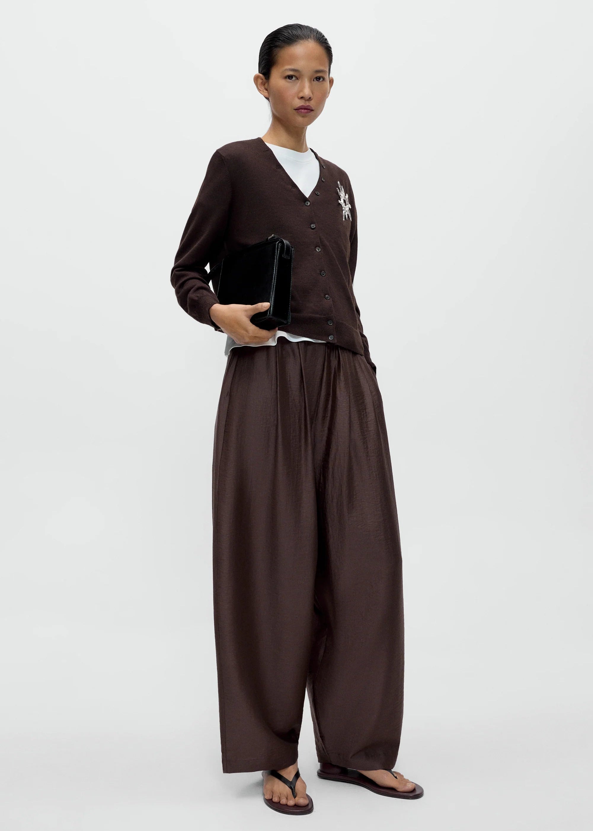 Person wearing a brown outfit holding a black clutch against a white background
