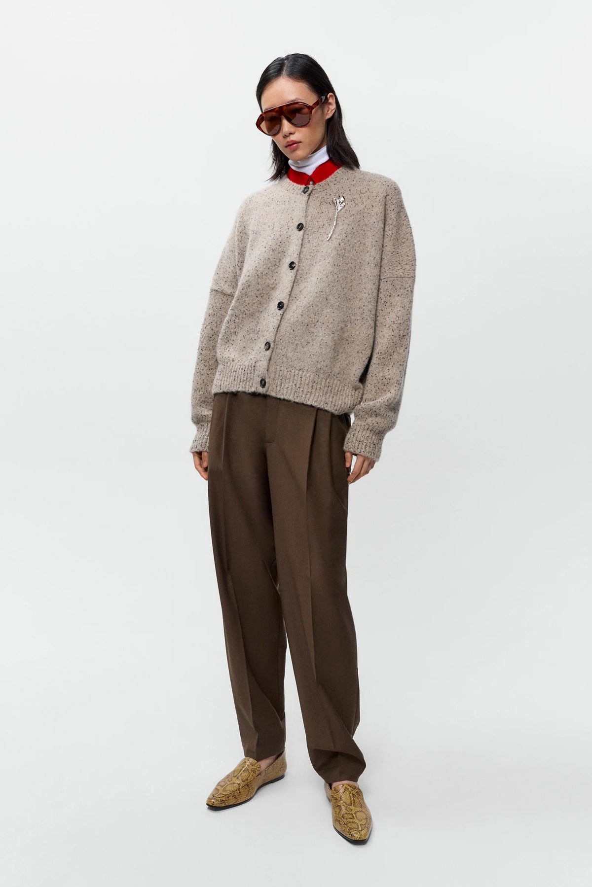 Person wearing a beige cardigan, brown pants, and sunglasses on a white background