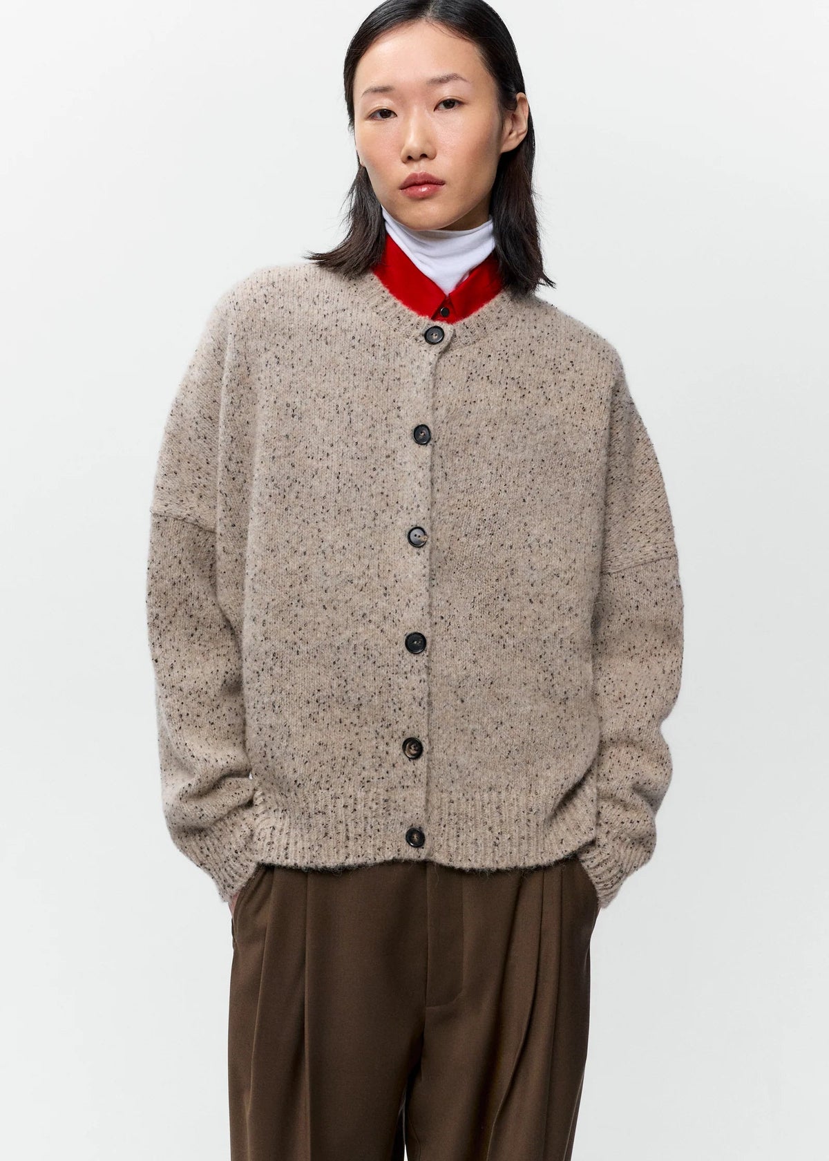 A model wearing a beige textured knit cardigan with a red collar, brown trousers, and a white turtleneck, photographed against a plain light gray background.
