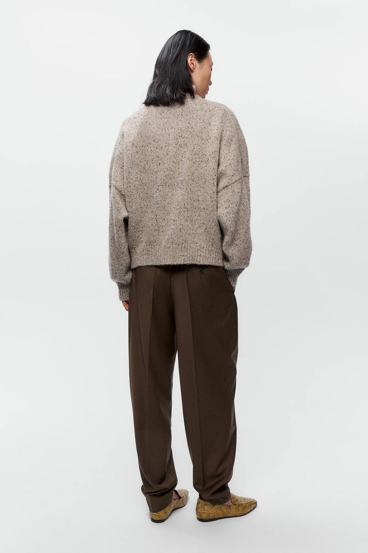 A model shown from the back wearing a textured, oversized beige knit sweater and dark brown wide-leg trousers, standing against a plain light gray studio background.