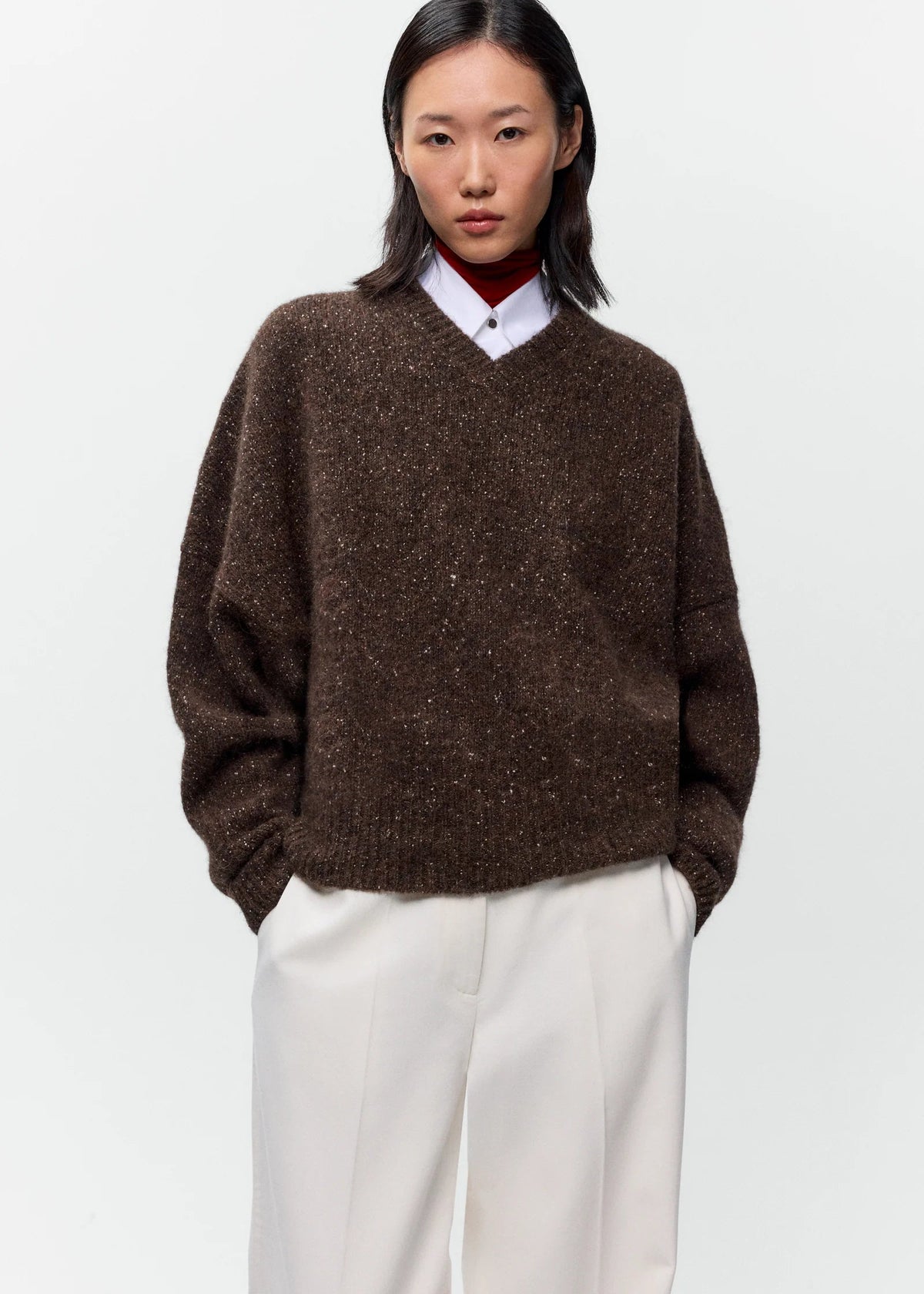 A model wearing a brown textured knit V-neck sweater over a white collared shirt and red turtleneck, paired with off-white trousers, against a plain light gray background.