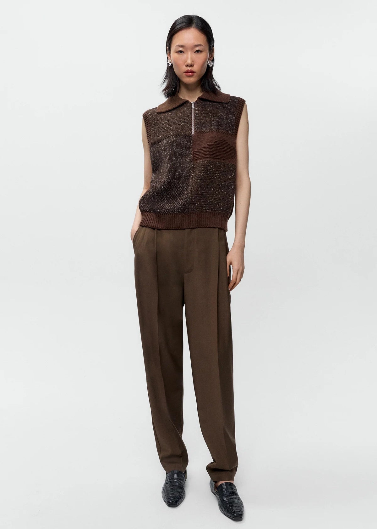 A model stands against a light gray studio background wearing a sleeveless brown knit vest with a mock neck and zipper, paired with matching brown trousers and black loafers.