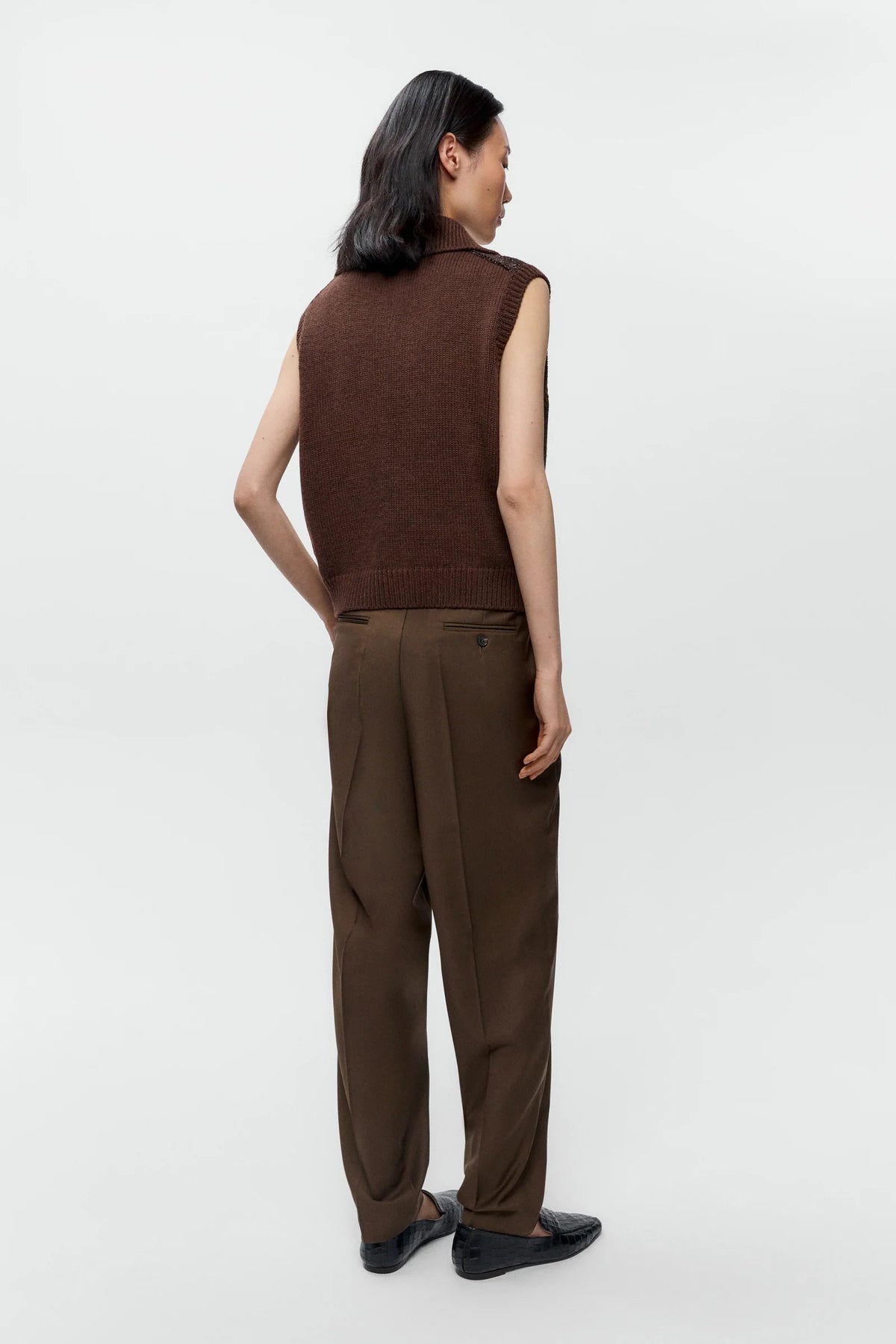 A model shown from the back wearing a brown sleeveless knit vest and matching brown trousers, paired with black loafers, against a plain light gray studio background.