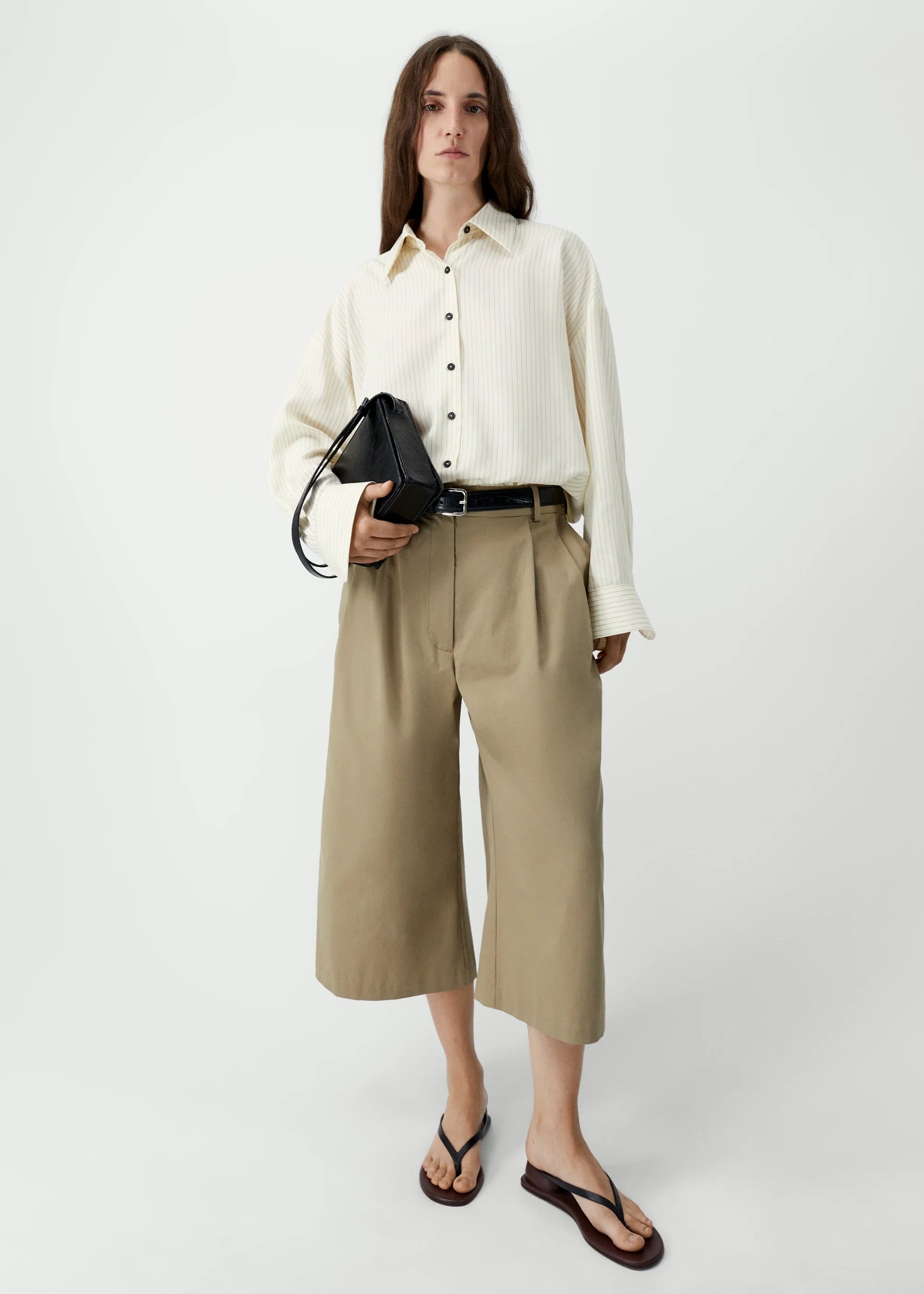 Woman wearing a white shirt and beige pants holding a black handbag on a white background