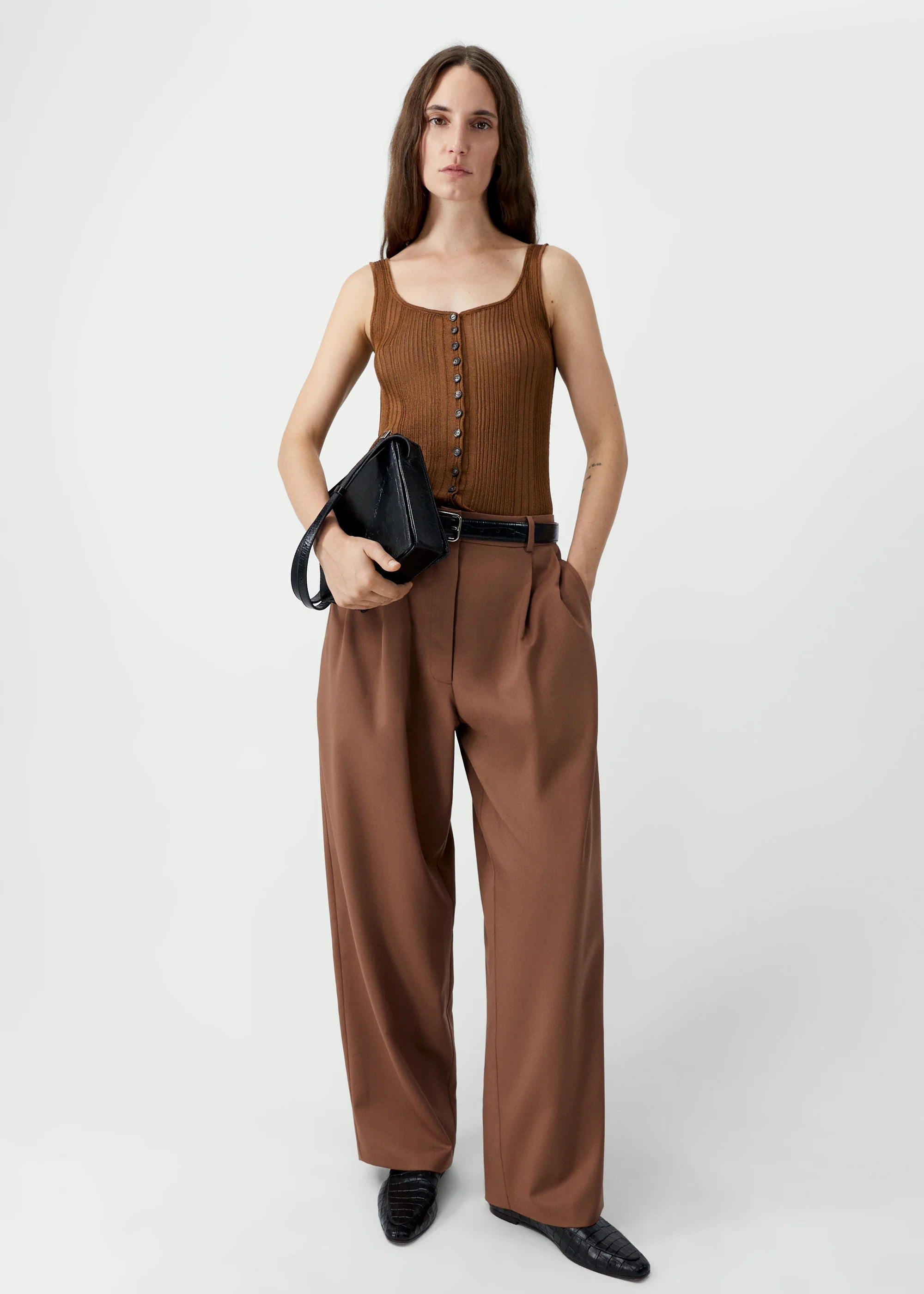 Woman wearing a brown sleeveless top and wide-leg pants holding a black clutch against a white background