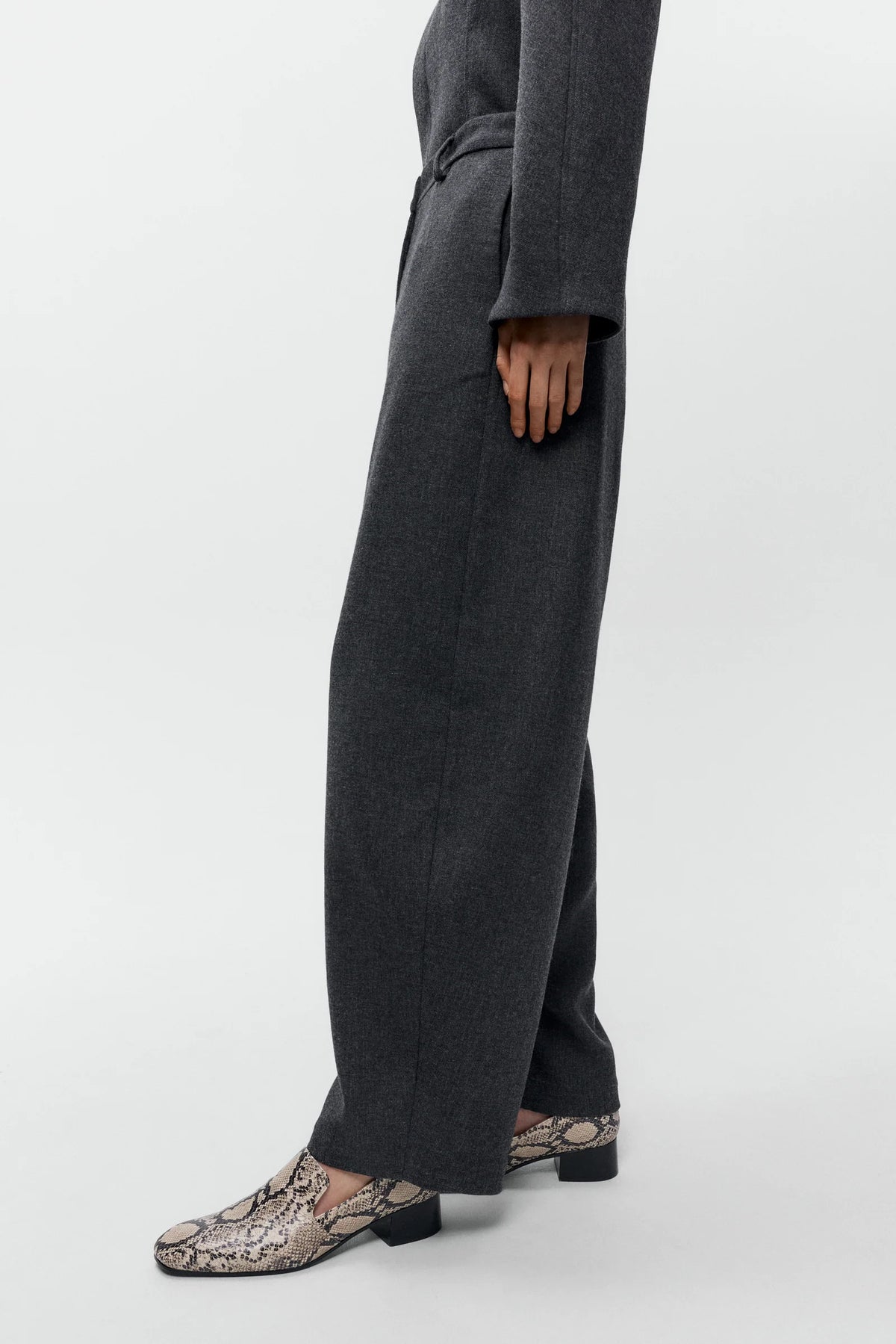 A person shown from the waist down wearing dark charcoal gray wide-leg trousers and snakeskin-patterned loafers, standing against a plain light gray studio background.