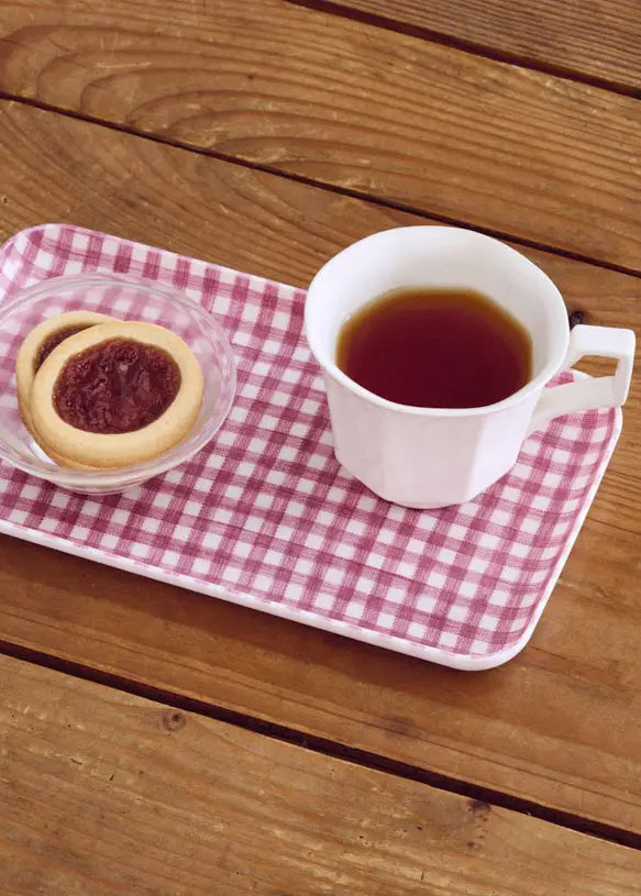 A rectangular tray with a pink and white gingham check pattern, holding a small clear bowl of cookies with jam and a white cup of tea, set on a rustic wooden table.