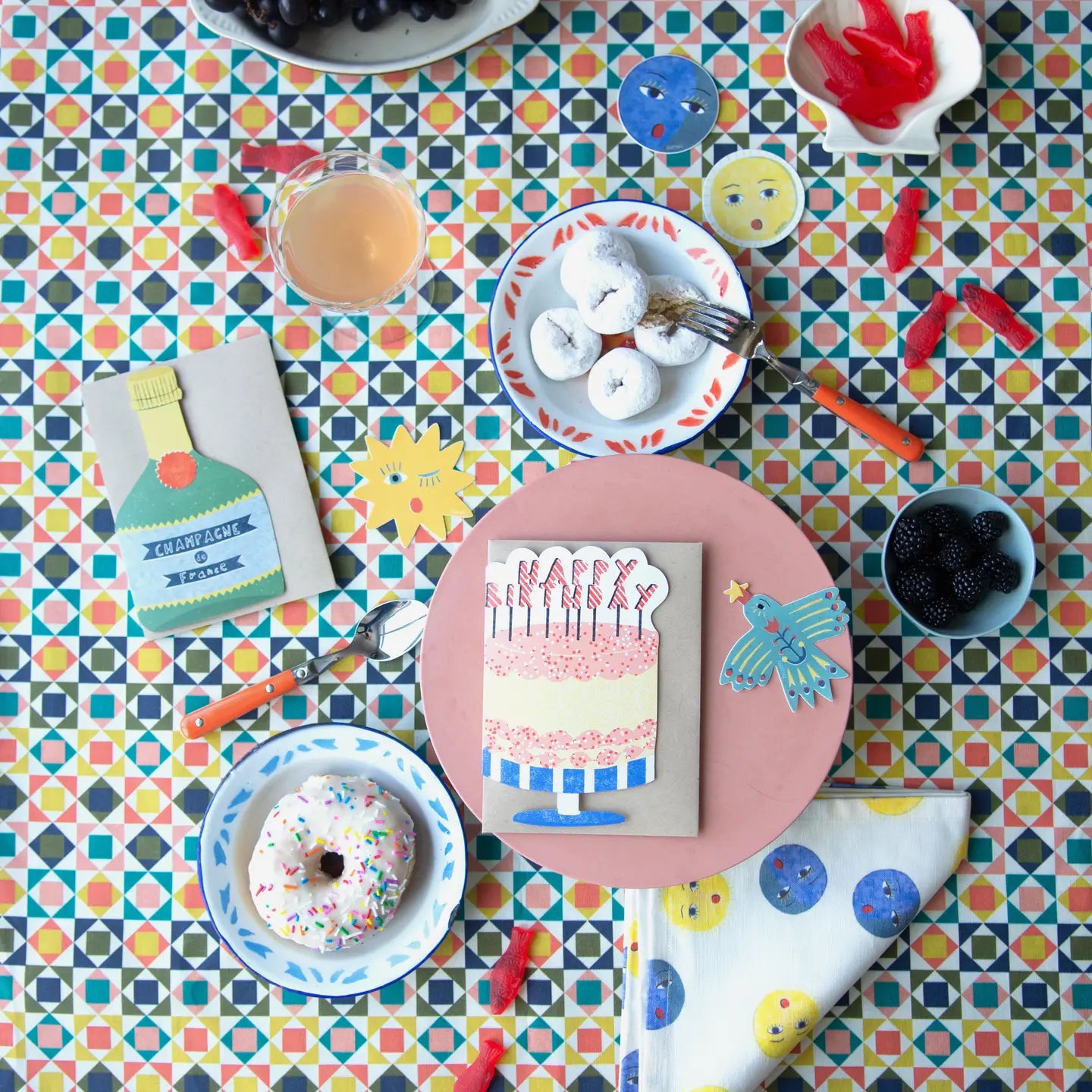 A colorful birthday party tabletop setup with a patterned multicolor tablecloth, a pink plate holding a birthday card that says 'Happy Birthday' with a cake illustration, a donut on a blue plate, white donuts on a plate with a fork, and small party accessories scattered around.