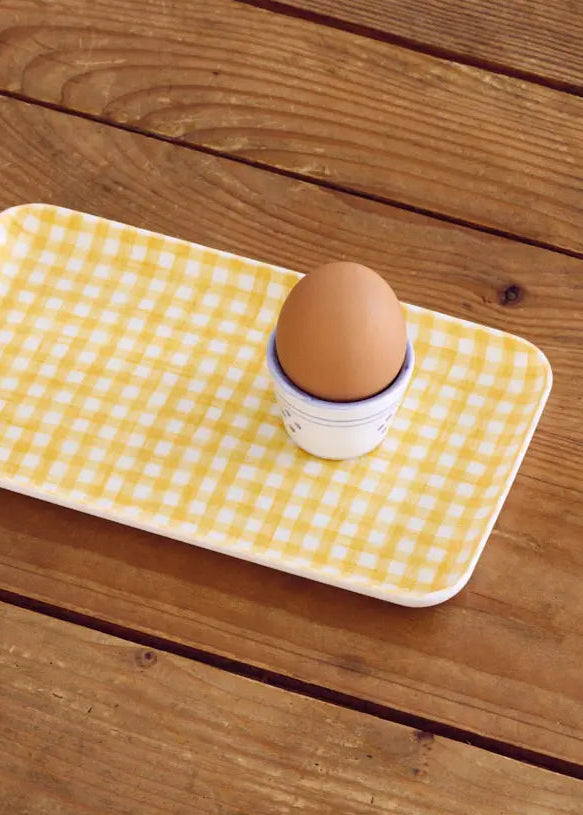 A rectangular white tray with a yellow and white gingham check pattern, holding a brown egg in a small white cup, set on a rustic wooden table.