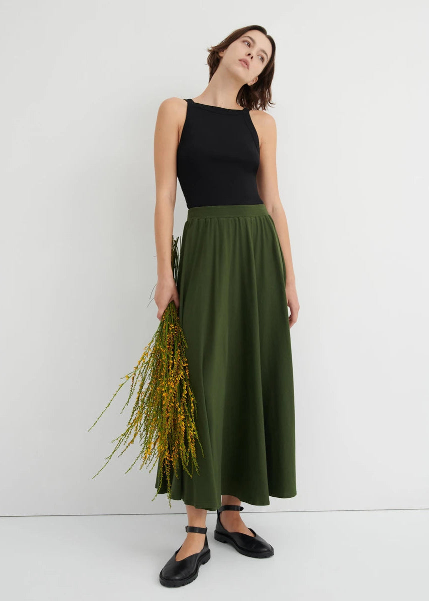 model wearing long green skirt with black tank and black shoes holding flowers
