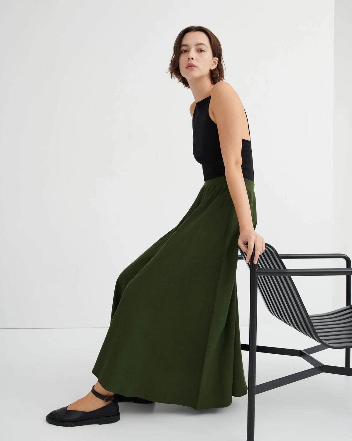 model wearing long green skirt with black tank and black shoes leaning on a chair